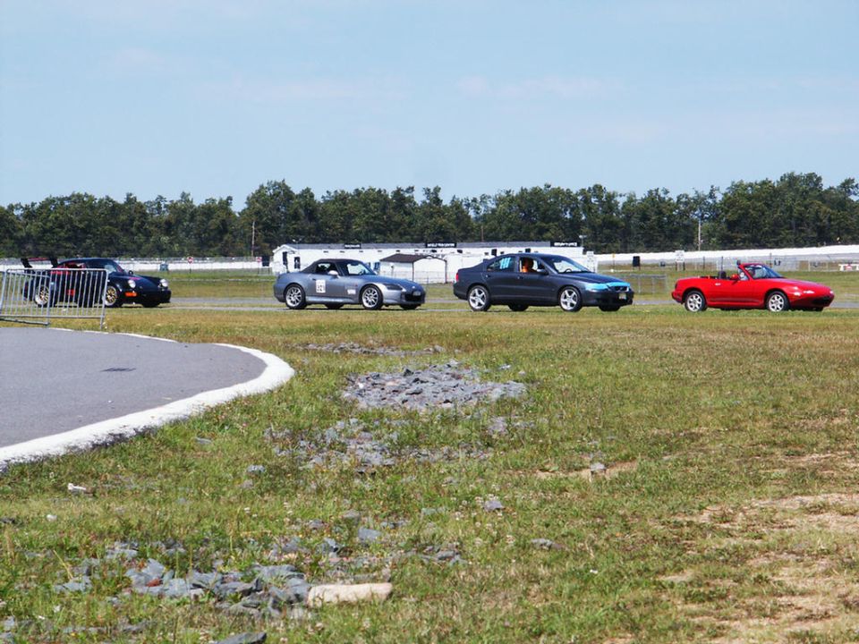 Four cars on a race track: a black Porsche, a silver Honda S2000, a dark sedan, and a red convertible