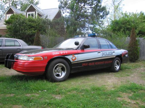 1991 Ford LTD Crown Victoria - A police car with a push bar and emergency lights is parked in a grassy area next to a house.