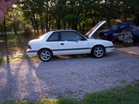 1990 Dodge Shadow - A white coupe sits in a gravel lot with its hood open, surrounded by trees and other vehicles.