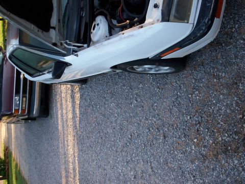 1990 Dodge Shadow - A white car with its hood open is parked next to another vehicle on a gravel surface.