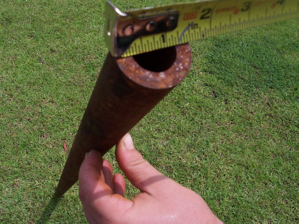 Rusty metal pipe being measured by a yellow tape measure against a grassy background