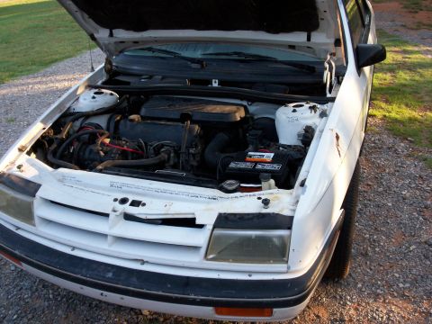 1990 Dodge Shadow - The engine bay of a white 1990 Dodge Shadow is visible with the hood open, revealing the engine and front grille.