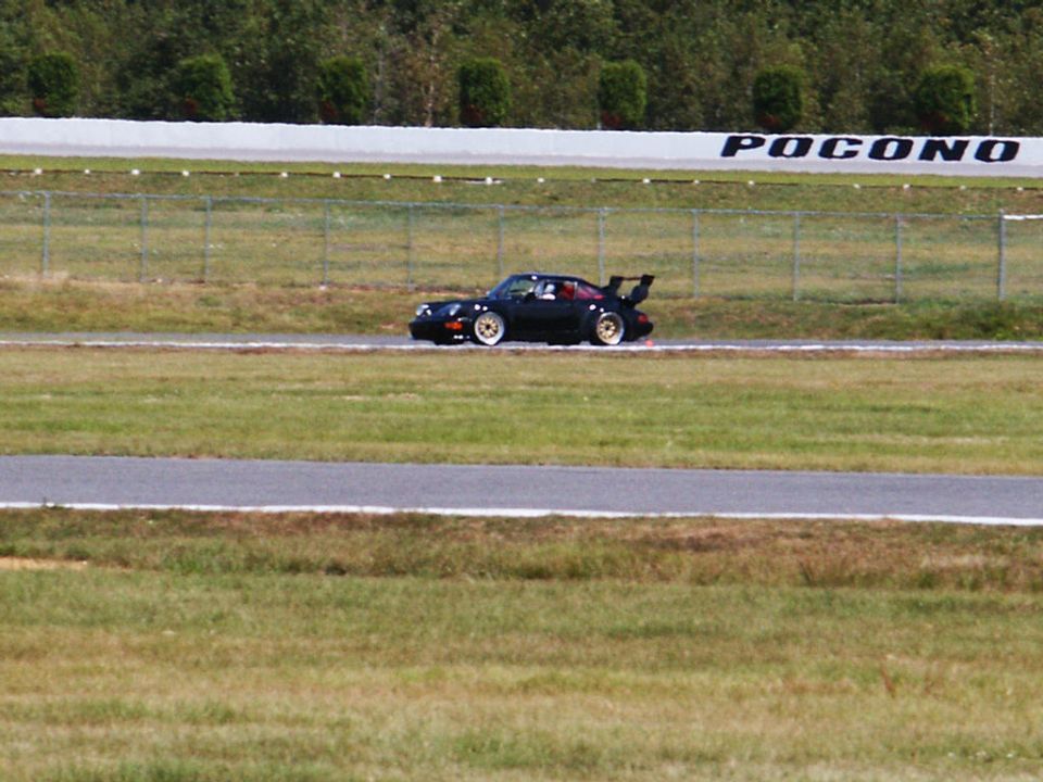 Black Porsche 911 with a large rear spoiler on a race track, with 'POCONO' visible on a sign in the background
