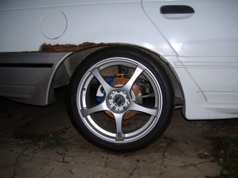 1998 Ford Taurus - Rear wheel and rusted quarter panel of a white sedan with aftermarket rims and blue brake calipers.