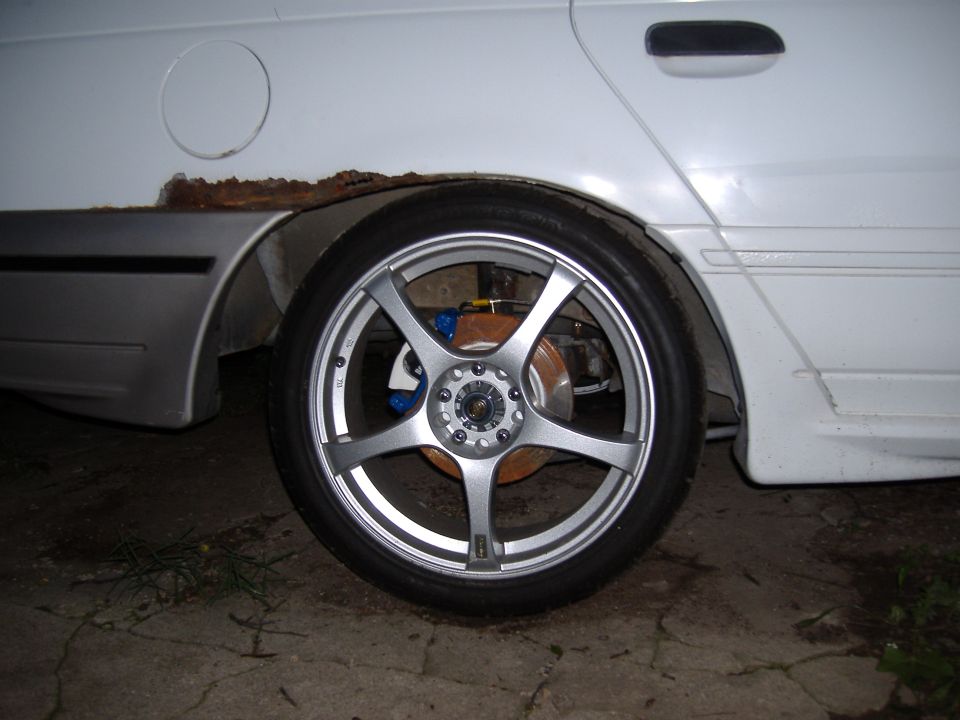 White sedan rear wheel with aftermarket rims and blue brake calipers, showing rust on the quarter panel