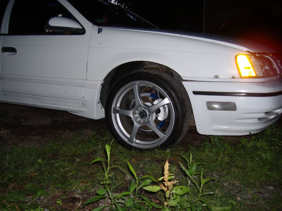 Front corner of a white sedan with aftermarket wheels and blue brake calipers. Turn signal is on