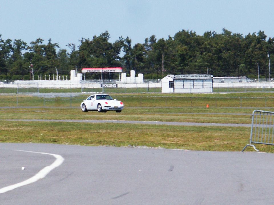 White sports car with racing number 21 on the side, driving on a grassy area next to a racetrack. A building labeled 'EVENT MERCHANDISE' is in the background