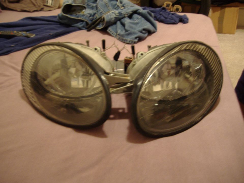 Pair of car headlights, possibly for a sedan, lying on a bed with clothes in the background