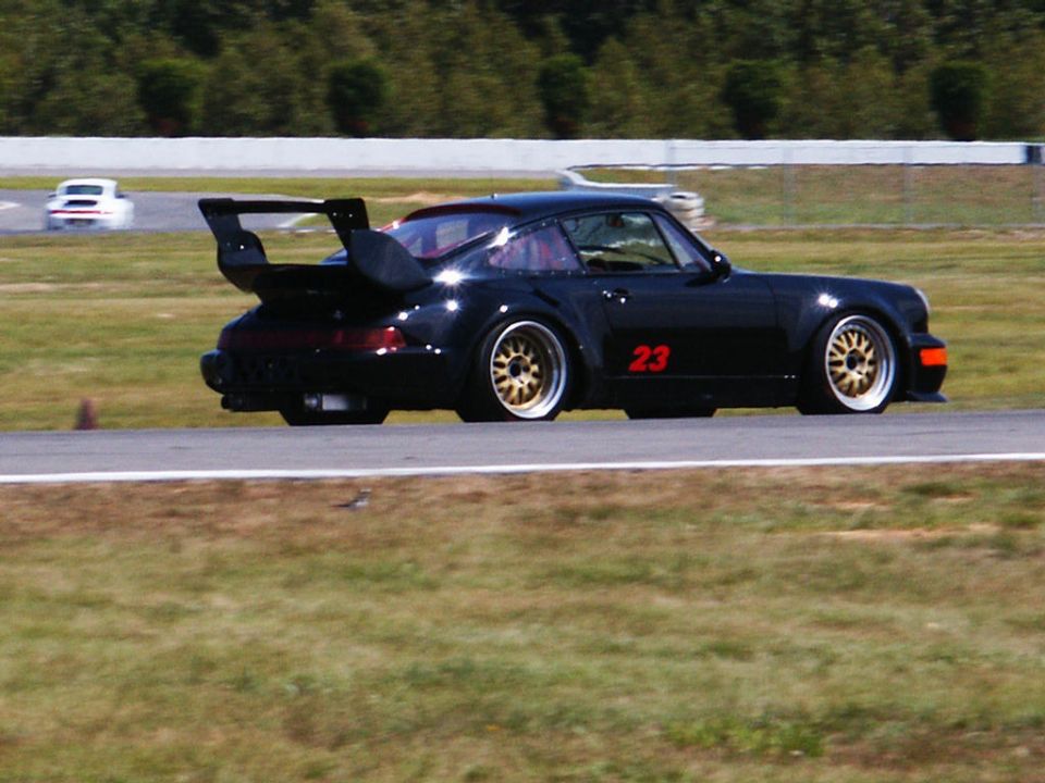 Black Porsche 911 with a large rear spoiler and gold wheels on a race track, another white car in the background