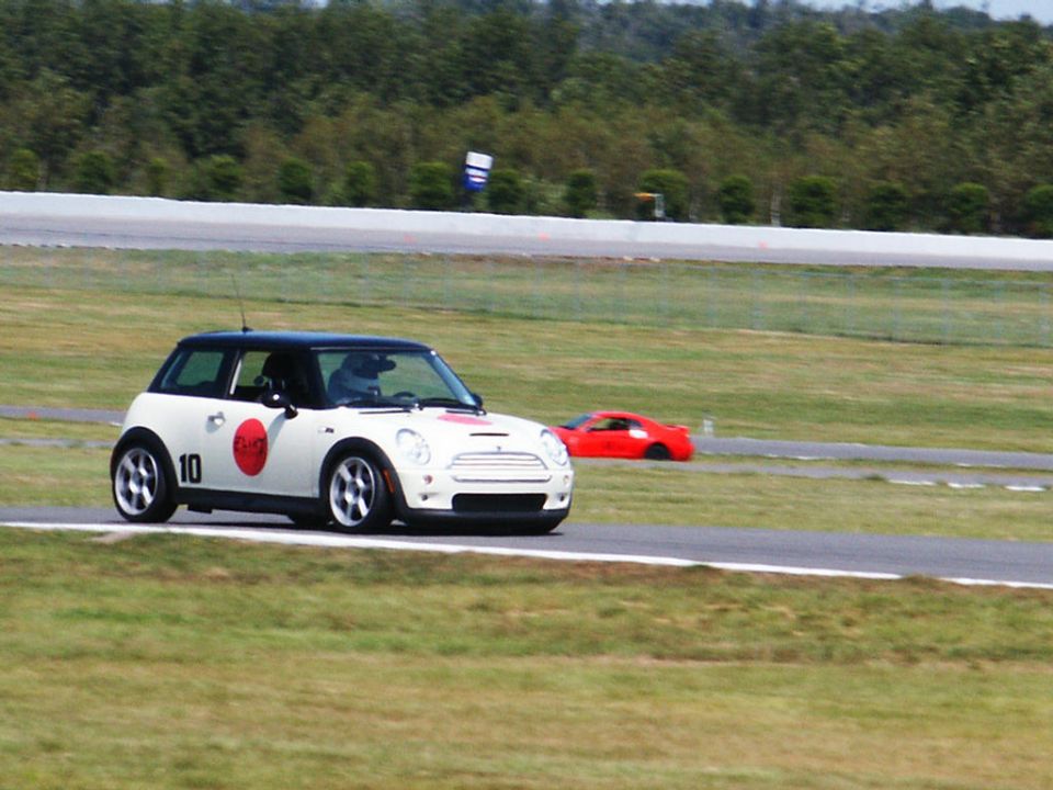 White Mini Cooper with red circle and number 10 on the side, on a race track. Red sports car in the background