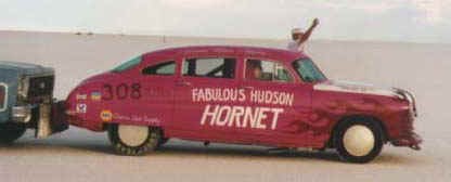 1970 Chevrolet Camaro - Pink 1950s Hudson Hornet drag car numbered 308 is seen towing another vehicle on a flat, sandy surface.