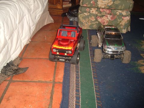 1988 Ford Bronco - Two remote control monster trucks are parked on a rug next to a bed.