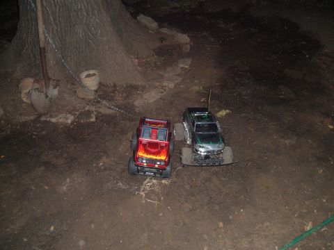 1988 Ford Bronco - Two remote control monster trucks are parked on a dirt path next to a large tree.