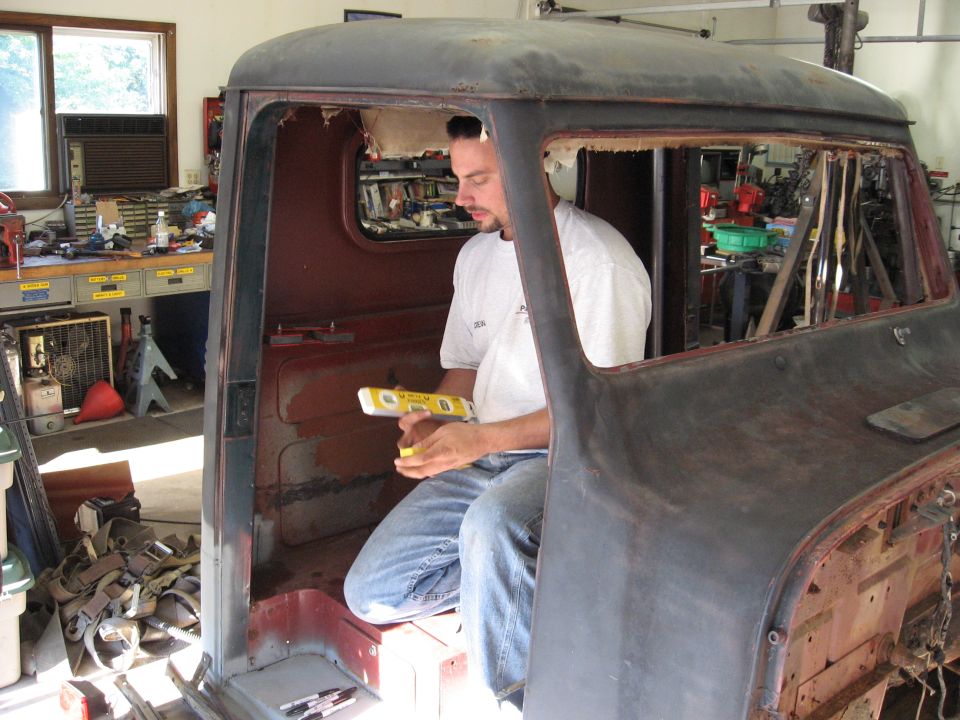 Man working inside a rusty, disassembled Ford truck cab, holding a yellow level