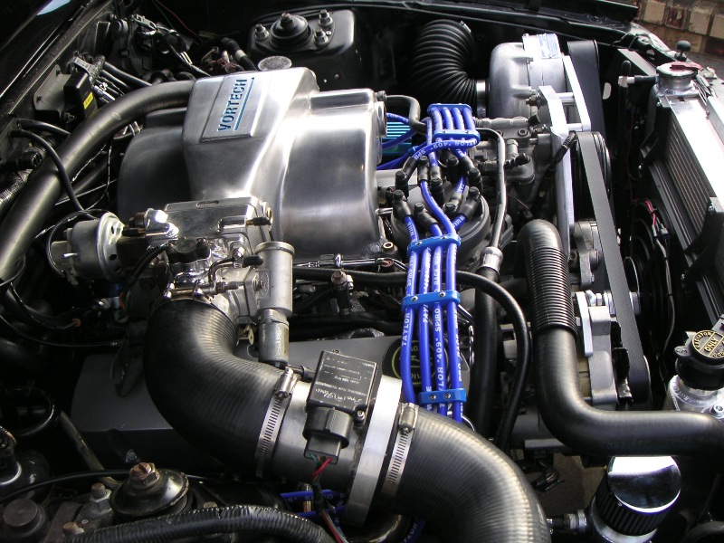 Engine bay with a silver Vortech supercharger, blue spark plug wires, and black hoses