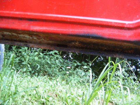 1993 Ford Bronco - Underside of a red vehicle, showing rust and dirt, with dense green foliage in the background.