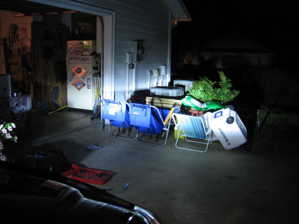 Nighttime driveway with coolers, lawn chairs and gear stacked by an open garage door, beer signs inside