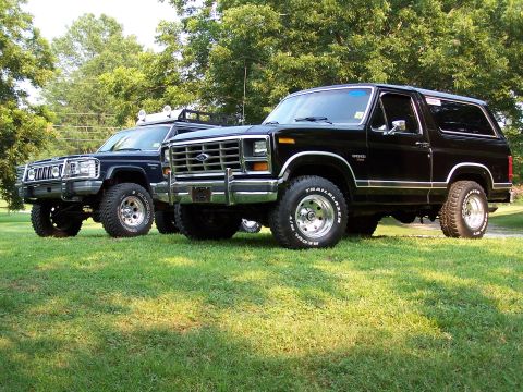 1983 Ford Bronco - Two off-road vehicles sit side-by-side on a grassy lawn under a canopy of trees.