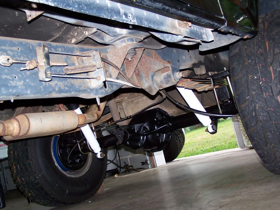 Underside view of a vehicle's rear axle, leaf springs, and exhaust system. New white shocks are visible