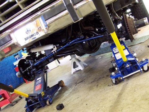 1983 Ford Bronco - Undercarriage view of a 1983 Ford Bronco during a suspension lift installation, supported by hydraulic jacks.