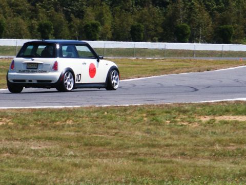 2001 Mercury Grand Marquis - A Mini Cooper with racing decals navigates a turn on a paved track surrounded by grass and trees.