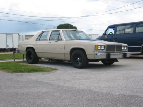 1985 Ford LTD Crown Victoria - A tan sedan with a blue hood accent sits parked on asphalt next to a dark blue van.