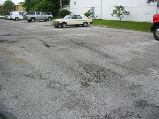 Cream-colored coupe parked in asphalt lot next to white van and silver SUV. Tire marks on pavement