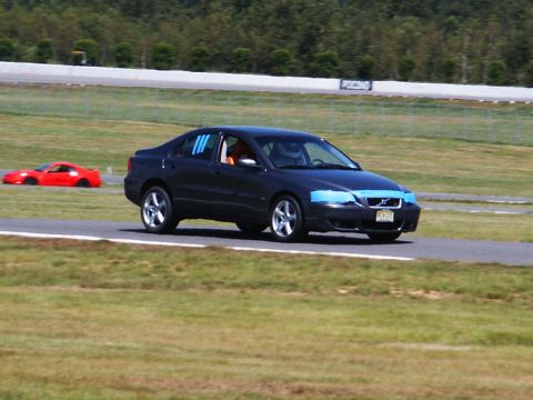 2001 Mercury Grand Marquis - A dark sedan and a red sports car are on a racetrack, with the sedan having blue tape on its headlights and blue stripes on its windows.