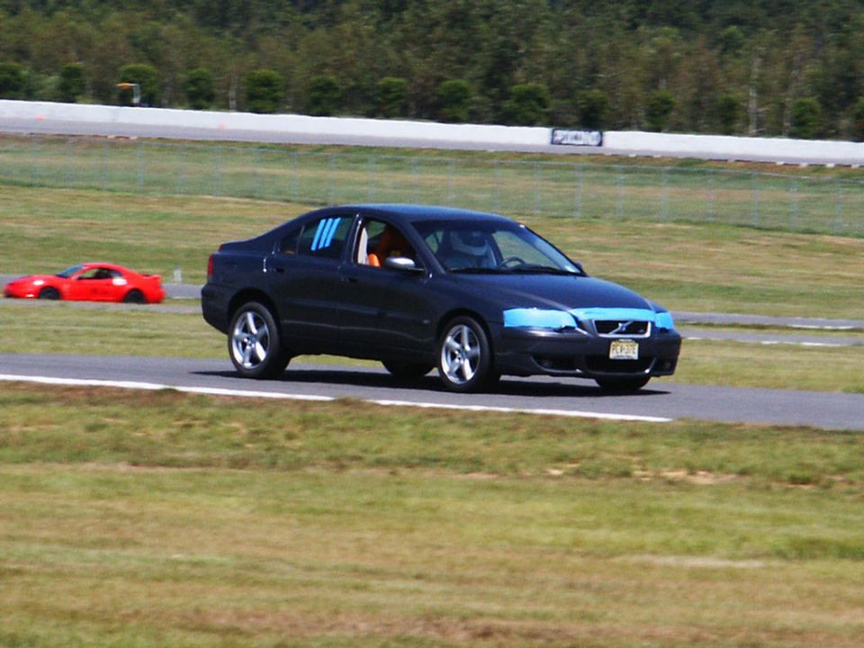 Dark sedan with blue tape on headlights and blue stripes on side windows drives on a racetrack next to a red sports car