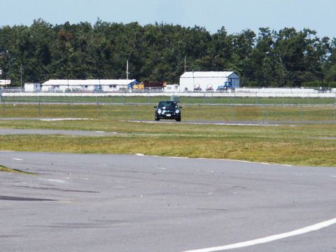 2001 Mercury Grand Marquis - A lone black car drives on a paved track with grassy areas and buildings in the background.