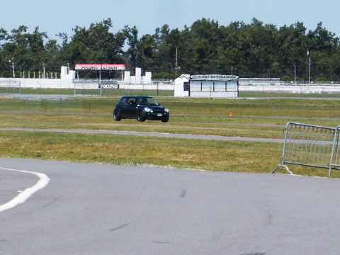 2001 Mercury Grand Marquis - A black Mini Cooper drives on the track at Pocono Raceway, with event merchandise stands visible in the background.