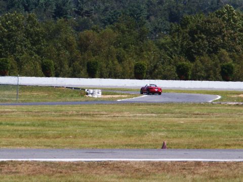 2001 Mercury Grand Marquis - A red convertible sports car navigates a turn on a race track, with trees and a white barrier in the background.