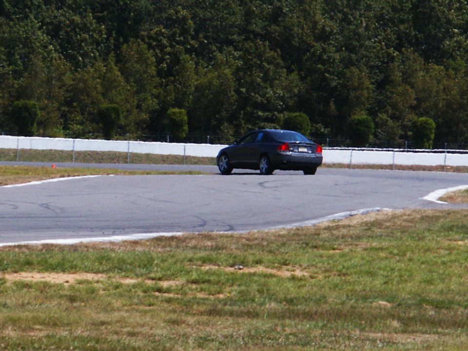 Dark sedan driving on a race track with trees and a white fence in the background