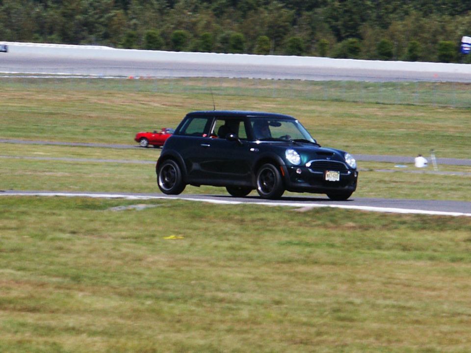 Dark green Mini Cooper on a race track with a red convertible in the background