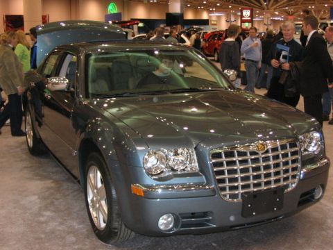 1994 Mazda 626 - A gray Chrysler 300 sedan is displayed at an auto show, with its hood open and people milling around.