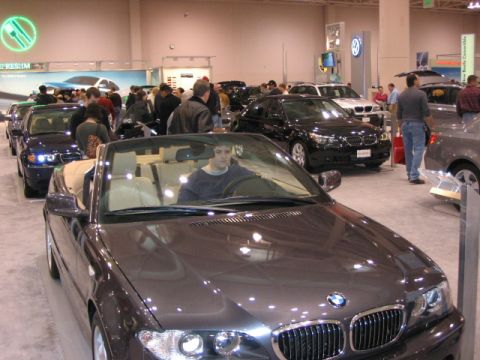 1994 Mazda 626 - A BMW convertible is displayed at an auto show, with other vehicles and attendees in the background.