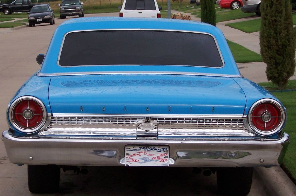 Rear of a blue 1963 Ford Galaxie with chrome bumper and round taillights. Other cars and a white SUV are visible in the background