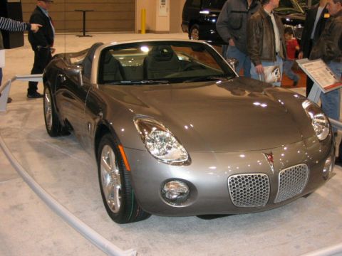 1994 Mazda 626 - A silver convertible sports car is displayed at an auto show, with people gathered around.