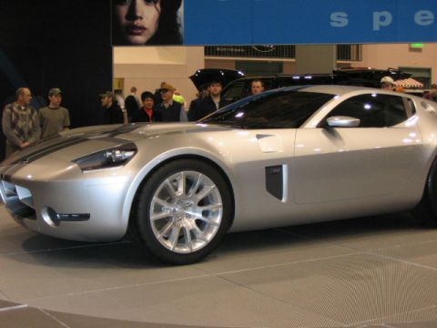 1994 Mazda 626 - A silver sports car with racing stripes is displayed at an auto show, with people in the background.