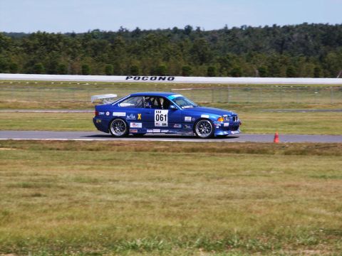 2001 Mercury Grand Marquis - A blue BMW race car is on the track at Pocono Raceway, with trees and a white barrier in the background.
