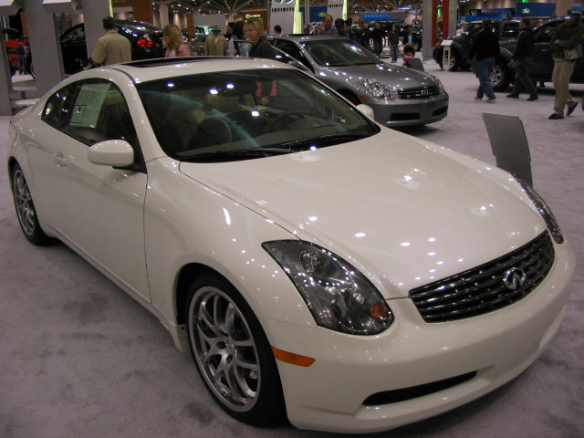 White coupe with sunroof and alloy wheels on display at an auto show, with a silver sedan behind it