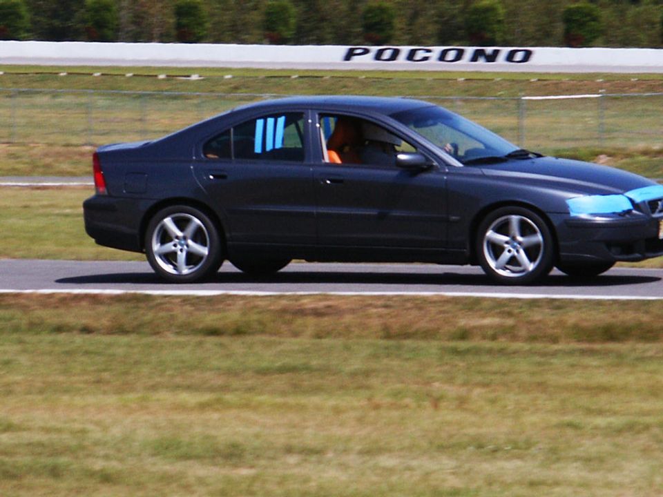Dark sedan with blue tape on headlights, driving on a track with 'Pocono' visible in the background