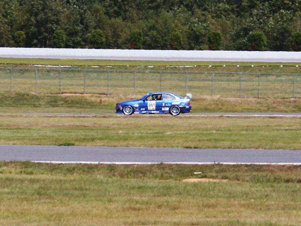Blue race car with number 061 on side, driving on a track with trees and fence in background