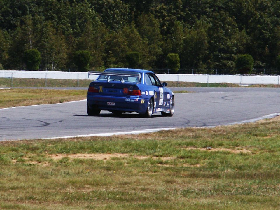 Blue race car with a spoiler on a track, seen from the rear. It has racing decals and the number 'X'