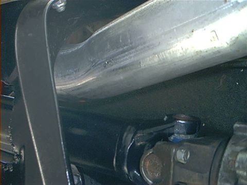 1978 Ford Bronco - Underbody close-up of a hand-formed stainless exhaust pipe with a pinched, welded bend clearing the black driveshaft and U-joint below it.…