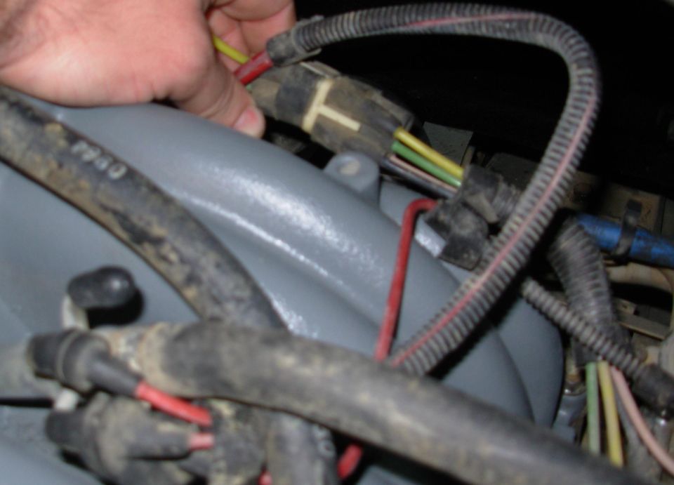 Hand holding wiring near a gray-painted engine bay component, frayed loom and red/yellow wires