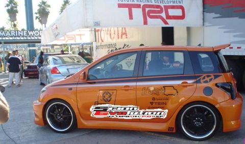 1993 Ford Taurus - An orange Scion xB is parked next to a silver sedan, with a Toyota TRD banner overhead.