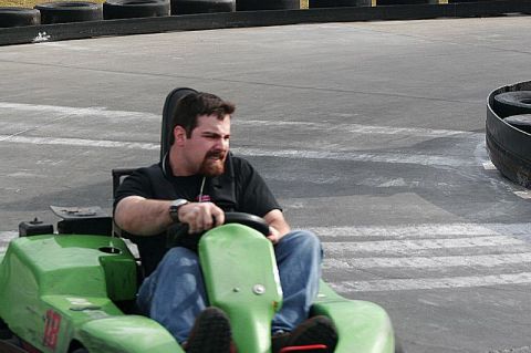1991 Ford Probe - A bearded driver in a black T-shirt and jeans leans into a turn aboard a green rental go-kart numbered 18, running on a concrete track line…
