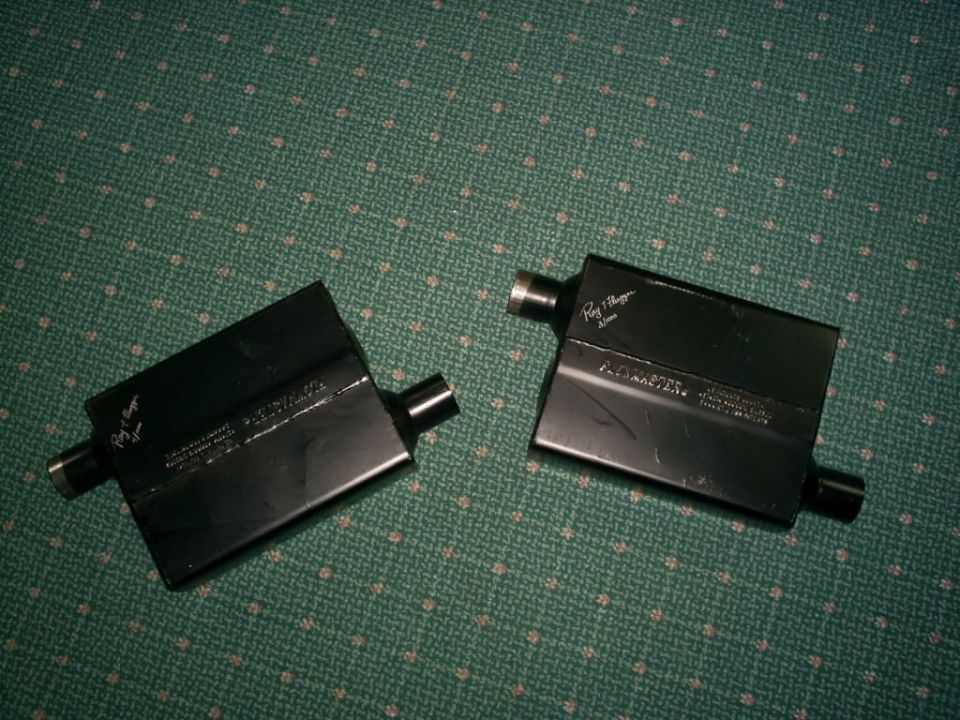 Two black exhaust mufflers with "Flowmaster" and "Ray J. Blagg" text visible, on a green patterned surface