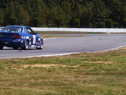 2001 Mercury Grand Marquis - A blue race car navigates a turn on a paved track with grassy shoulders.
